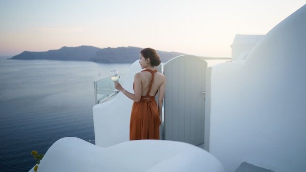 Mulher turista em um lindo vestido com uma taça de vinho relaxante ao pôr do sol