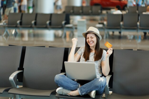 Mulher turista viajante alegre com laptop sentada com as pernas cruzadas, gesto vencedor, segurar cartão de crédito, esperar no saguão do aeroporto