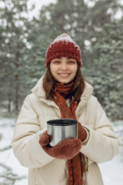 Mulher turva com garrafa térmica de chá quente em pé na floresta de neve no inverno e aquecendo as mãos