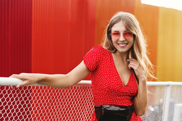 Mulher urbana com vestido de verão e óculos escuros, em pé perto de uma parede vermelha ao ar livre.