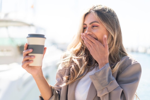 Mulher uruguaia muito loira segurando um café para viagem ao ar livre com surpresa e expressão facial chocada