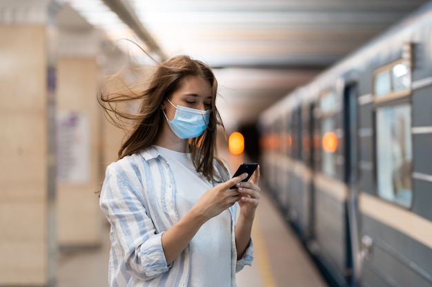 Mulher usa máscara facial usando celular, conversando nas redes sociais esperando o trem do metrô na plataforma