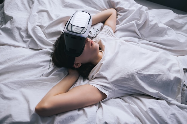 Mulher usa óculos de realidade virtual em casa, relaxada na cama. Joga jogos, tecnologias do futuro