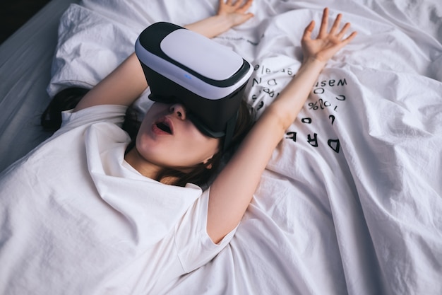 Mulher usa óculos de realidade virtual em casa, relaxada na cama. Joga jogos, tecnologias do futuro