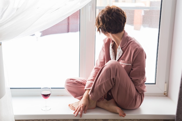 Mulher usa pijama com uma taça de vinho tinto sentada no parapeito da janela perto da bela janela em casa.