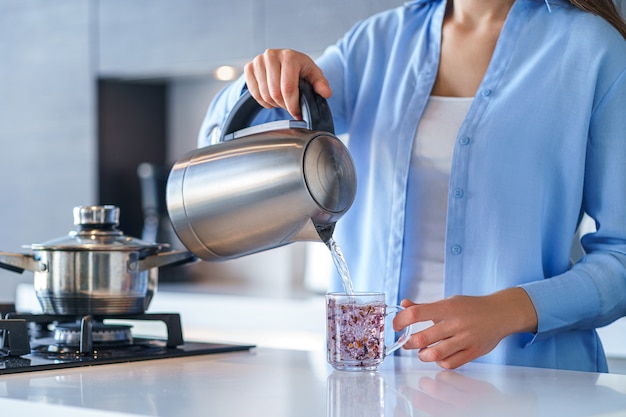 Mulher usando a chaleira elétrica de metal prateado para ferver água e fazer chá em casa. Utensílios de cozinha para bebidas quentes