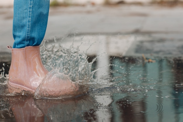 Mulher usando botas de borracha de chuva andando correndo e pulando em uma poça com respingos de água e gotas de chuva de outono.