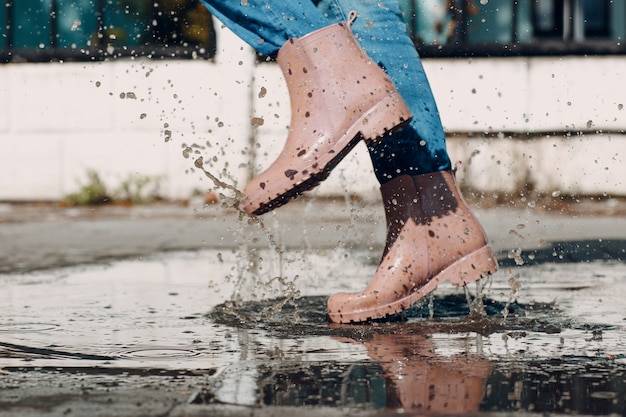 Mulher usando botas de borracha de chuva andando correndo e pulando em uma poça com respingos de água e gotas ...