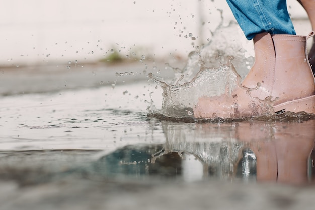 Mulher usando botas de borracha de chuva caminhando correndo e pulando em uma poça com respingos de água e gotas de chuva de outono.