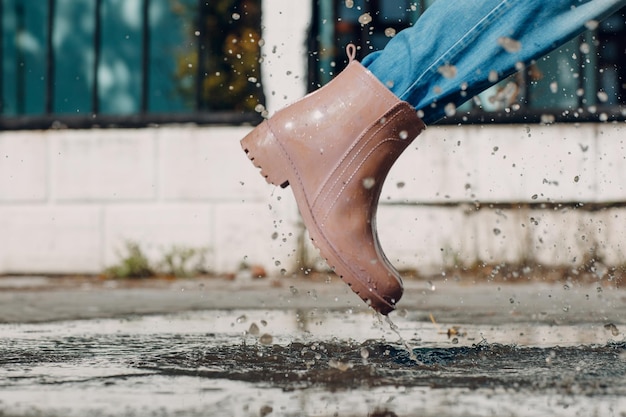 Mulher usando botas de borracha de chuva caminhando correndo e pulando em uma poça com respingos de água e gotas de chuva de outono.