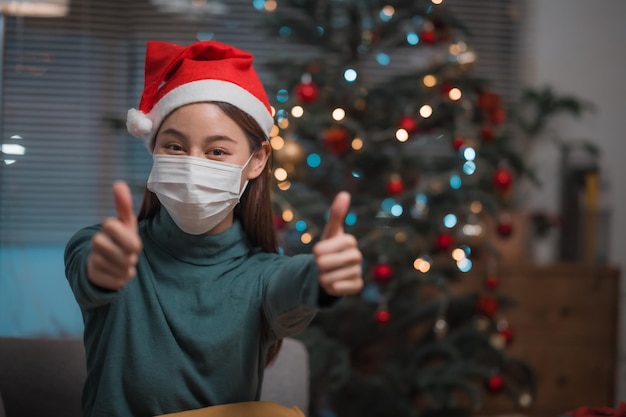 Mulher usando chapéu de Papai Noel e máscara protetora enquanto faz sinal de positivo durante o surto do vírus corona