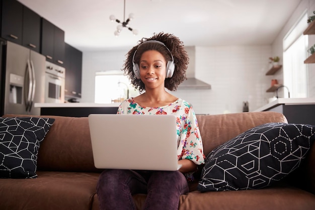Mulher usando fones de ouvido sem fio sentado no sofá em casa usando laptop