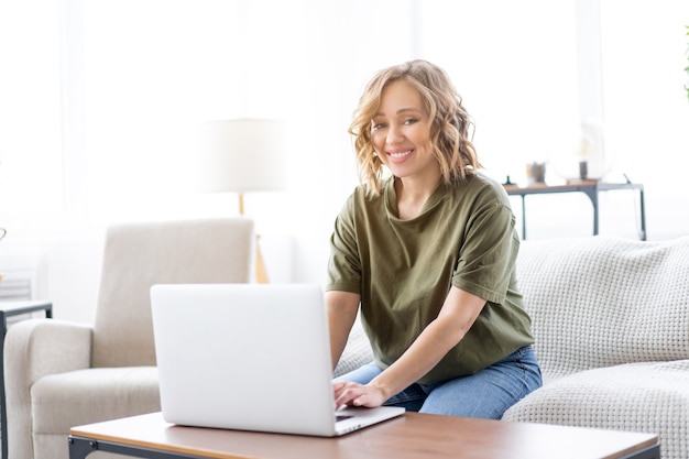 Mulher usando laptop enquanto está sentada no sofá janela grande fundo interior da casa Mulher autônoma trabalhando em casa