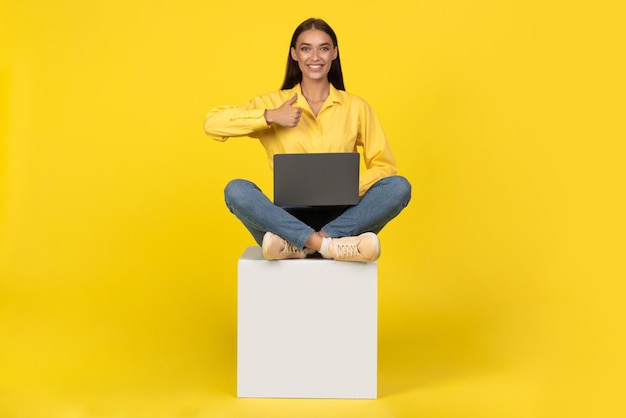 Mulher usando laptop gesticulando polegares para cima sentado sobre fundo amarelo