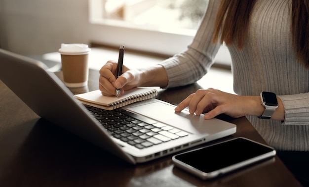 Mulher usando laptop para trabalhar e escrevendo no caderno com caneta na mesa com celular ao lado do laptop