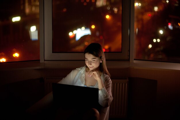 Mulher usando laptop sentada perto da janela em casa na cena da cidade à noite