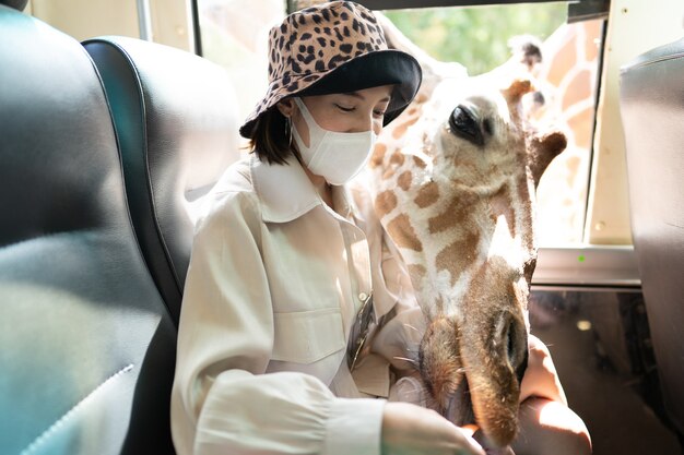 Mulher usando máscara com girafa esperando por comida na janela do ônibus no zoológico