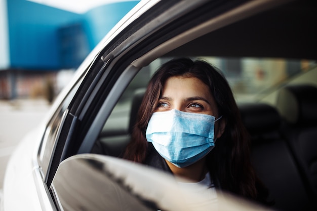Mulher usando máscara médica estéril em um carro de táxi no banco de trás, olhando de lado pela janela aberta