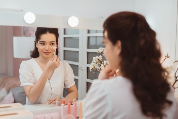 Mulher usando rolo facial de jade para cuidados com a pele, tratamento de beleza diário matinal perto da mesa