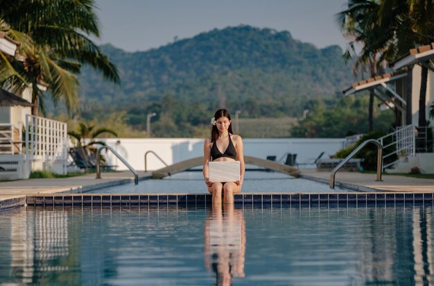 Mulher usando seu laptop na beira da piscina em um dia ensolarado Trabalho freelancer em país tropical