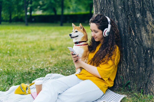 Mulher usando smartphone ouvindo música em fones de ouvido no parque verde com cachorro