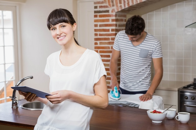 Mulher usando tablet na cozinha enquanto homem passando uma camisa