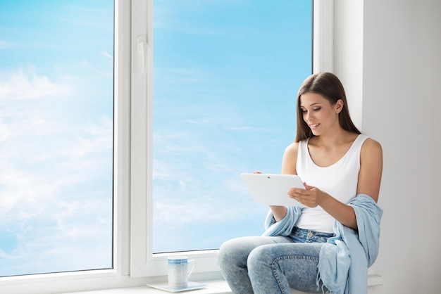 Mulher usando Tablet sentada no peitoril da janela em casa Garota lendo teclado digital branco Fundo do céu azul
