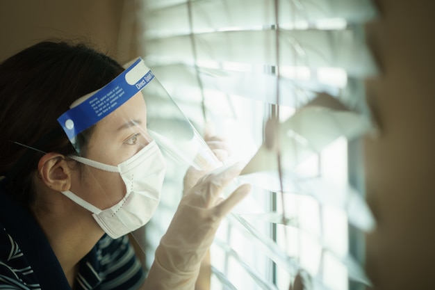 Mulher usando tela médica de proteção ou escudo plástico, olhando através das cortinas da janela. Para vírus corona ou proteção Covid-19