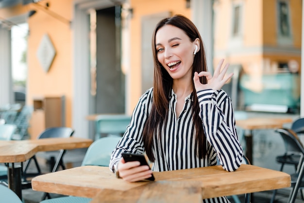 Mulher usando telefone e fones de ouvido sem fio sentado no café ao ar livre