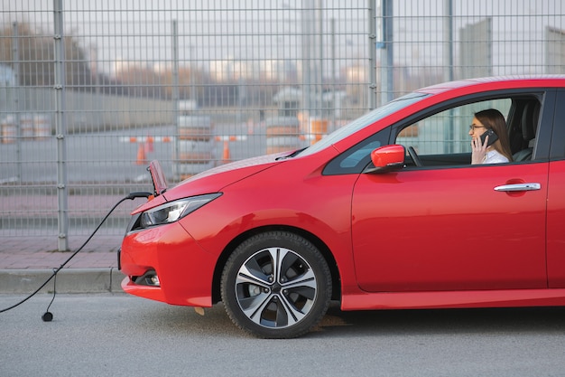 Mulher usando telefone inteligente e espera a fonte de alimentação conectar-se a veículos elétricos para carregar a bateria no carro. Jovem positiva falando ao telefone senta-se no carro elétrico e carregamento.