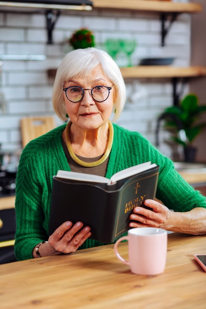 Mulher usando um elegante cardigã verde e óculos segurando um livro aberto da bíblia sagrada