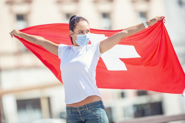 Mulher usando uma máscara facial está de pé com os braços abertos ao ar livre, segurando uma bandeira da Suíça atrás dela.