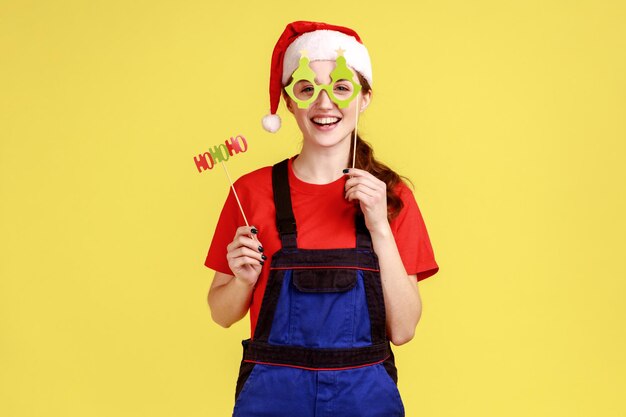 Mulher útil engraçada se divertindo, experimentando máscaras de festa de natal na vara, comemorando férias de inverno, vestindo macacão azul e chapéu de papai noel. Tiro de estúdio interior isolado em fundo amarelo.