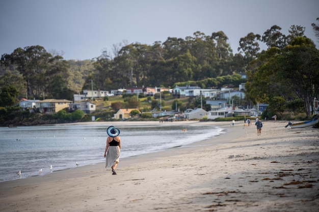 Foto mulher velha caminhando na praia de areia ao lado do mar com ondas quebrando atrás e montanhas à distância