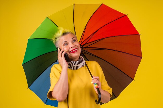 Mulher velha com cabelo tingido de amarelo elegante sob o guarda-chuva colorido do arco-íris segurando o telefone no fundo amarelo do estúdio.