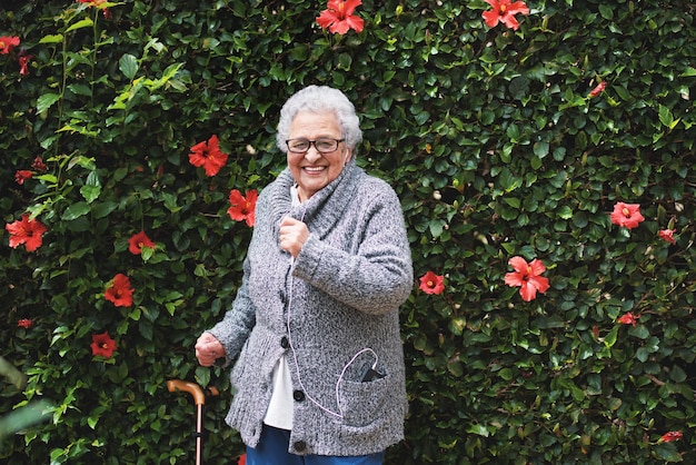 Mulher velha engraçada dançando ouvindo música no smartphone usando fones de ouvido sorrindo se divertindo comemorando a aposentadoria no jardim com parede de flores