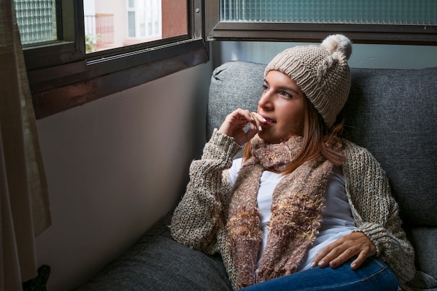 Mulher vestida com roupas quentes por causa do inverno frio, sentada no sofá ao lado da janela.