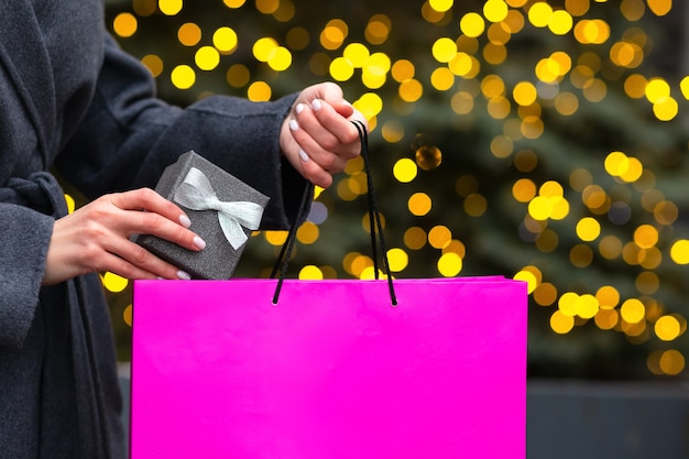 Mulher vestida com um casaco cinza tira uma caixa de presente com um laço da sacola de compras no fundo do bokeh e luzes desfocadas. Espaço vazio