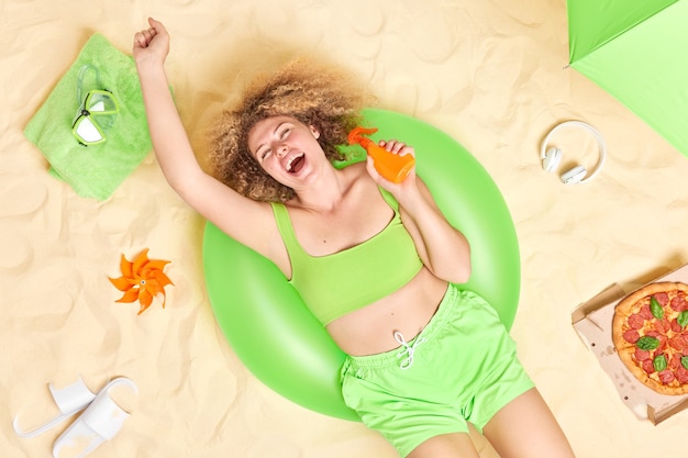 mulher vestida com uma blusa verde e shorts segurando uma garrafa de protetor solar posa na natação inflada passa o tempo livre na praia comendo pizza e tem um dia de preguiça