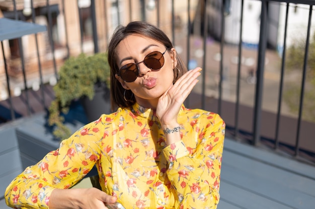 Mulher vestida de amarelo no terraço de um café de verão com telefone celular em um dia de sol, parece feliz e positiva manda beijo no ar