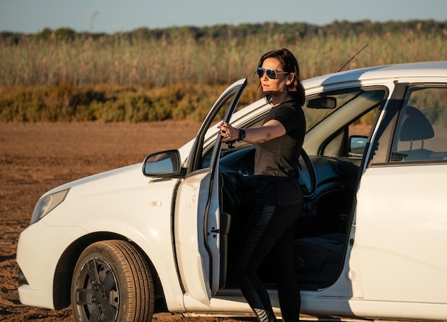 Mulher vestida de preto segurando a porta de um carro branco usando óculos escuros com o sol no rosto