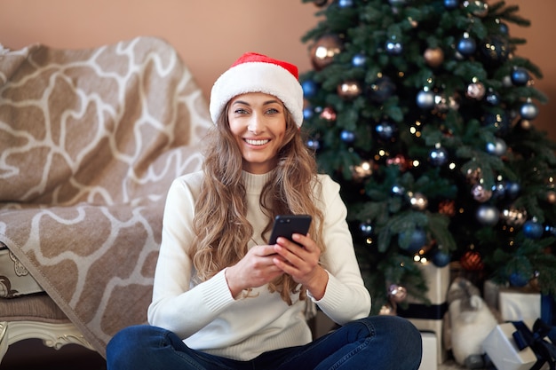 Mulher vestida de suéter branco com chapéu de Papai Noel no chão perto da árvore de Natal com smartpone e uma xícara de café