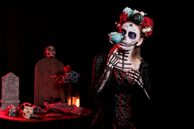 Mulher vestindo arte corporal para representar la cavalera catrina, posando com coroa de rosas e flores no estúdio. celebrando dios de los muertos com maquiagem de caveira e fantasia de deusa da morte.