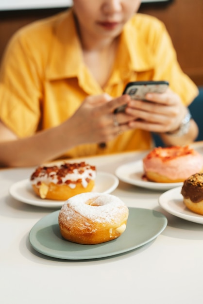 Foto mulher vestindo camisa amarela tirando fotos de morango rosa, chocolate, açúcar glaceado e bacon, donut de queijo.