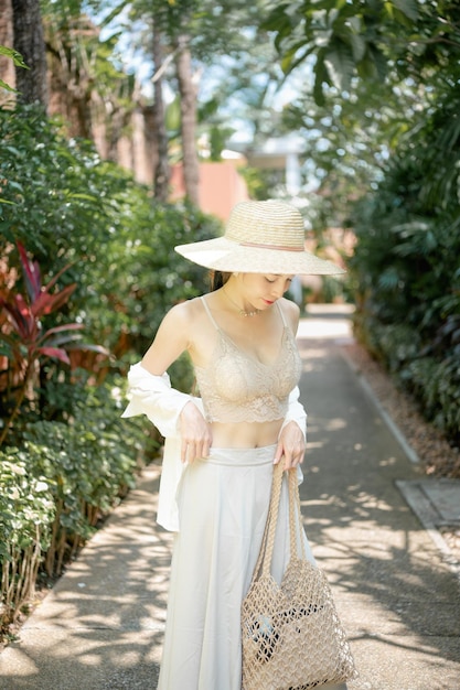 Mulher vestindo camisa branca, saia longa e chapéu de palha posando jardim tropical.