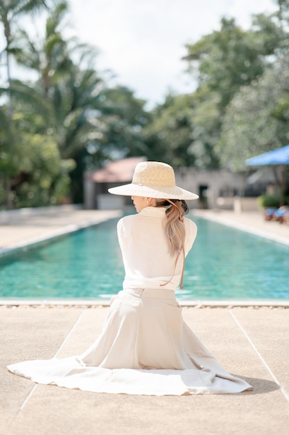 Mulher vestindo camisa branca, saia longa e chapéu de palha posando perto da piscina.