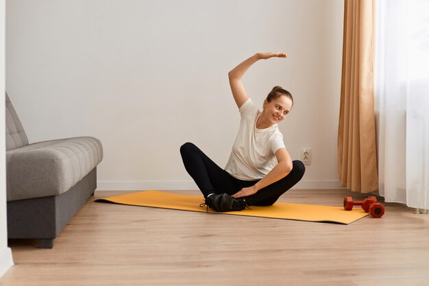 Mulher vestindo camiseta branca e leggins pretas fazendo exercícios esportivos em casa sentado em posição de lótus no tapete praticando ioga dobrando para o lado com a mão levantada fazendo exercícios de alongamento