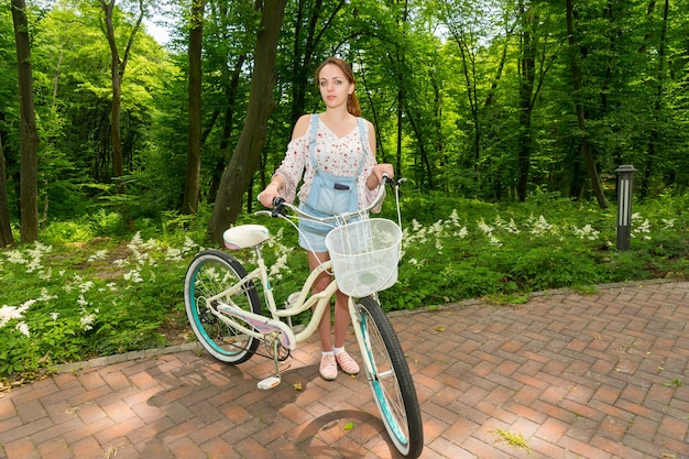 Mulher vestindo macacão jeans e uma blusa larga em pé com a bicicleta em tijolos em um parque