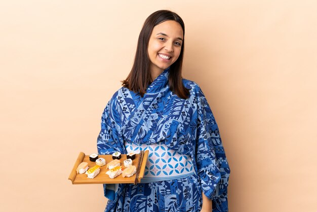 Mulher vestindo quimono e segurando sushi sobre parede isolada, olhando para o lado e sorrindo