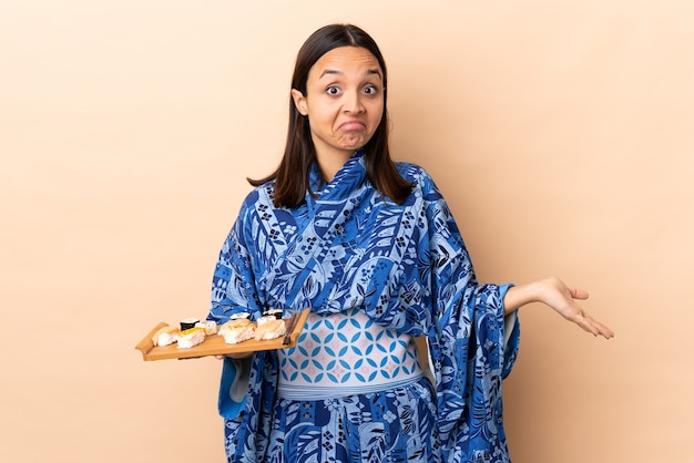 Mulher vestindo quimono e segurando sushi sobre um fundo isolado, tendo dúvidas enquanto levanta as mãos